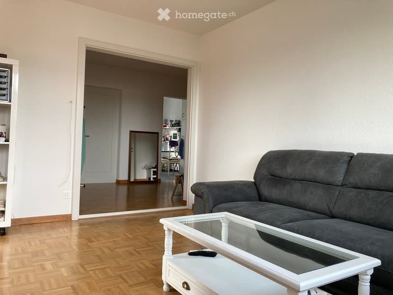 Living room with a grey sofa, a white coffee table, a mirror, and a shelf. The room has wooden flooring and white walls.
