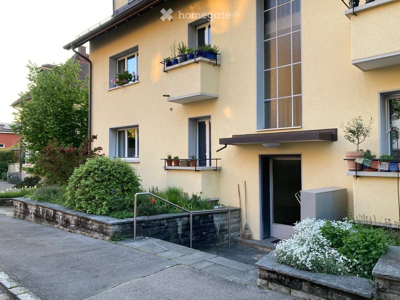 Two-story yellow building with multiple windows, balconies, and potted plants. Features a raised ground floor with a stone retaining wall and greenery.