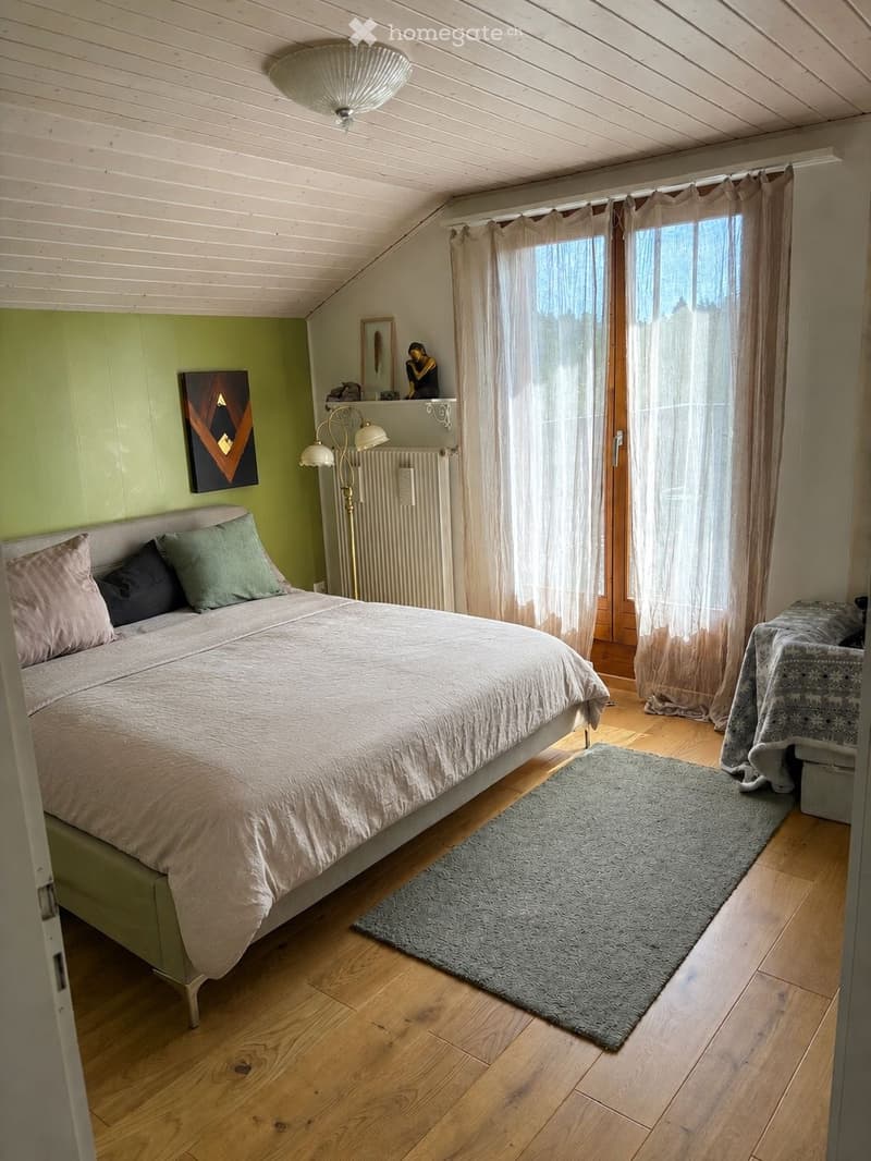 Cozy bedroom with wooden flooring, a large bed with pillows and a duvet, a nightstand with a lamp, a small table with a chair, and a window with sheer curtains. The room features a slanted wooden ceiling and a radiator under the window.