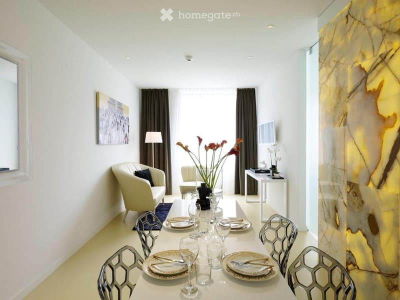 Modern dining room with a white dining table set for six, surrounded by geometric chairs, a cream sofa, a floor lamp, and a flower vase on a side table.
