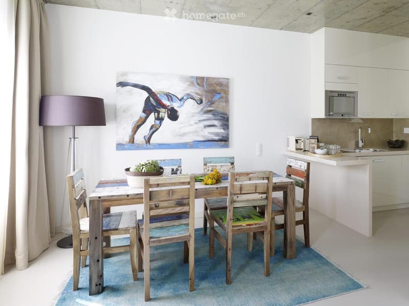 Open-plan dining and kitchen area with a wooden dining table, colorful chairs, a floor lamp, and modern kitchen cabinets.