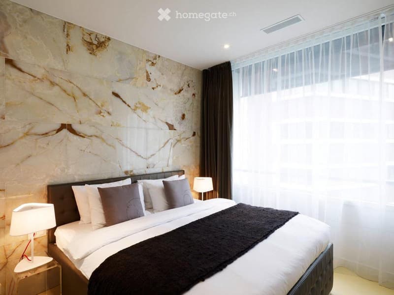 Modern bedroom with a large bed, decorative pillows, a dark throw blanket, and bedside lamps. The walls feature a marble-patterned design, and the room is illuminated by natural light from large windows with sheer curtains.