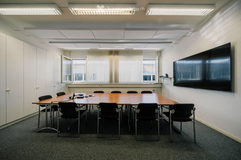Conference room with a large wooden table, black chairs, and a large flat-screen TV mounted on the wall. The room has white walls, carpeted flooring, and large windows with blinds.