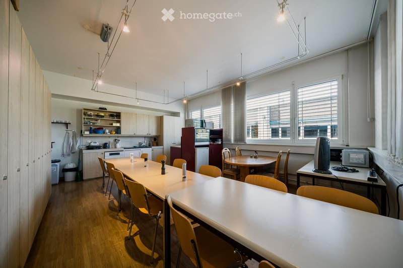 Bright, open-plan room with a long dining table and chairs, a smaller round table with chairs, a kitchen area with cabinets, a refrigerator, microwave, and a counter, and a workspace with a screen and other items.