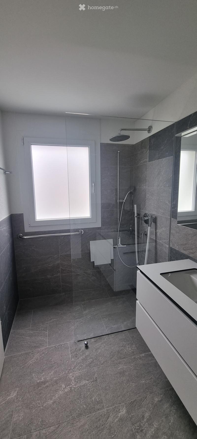 Modern bathroom with grey marble tiles, glass shower enclosure, white sink, and large window providing natural light.