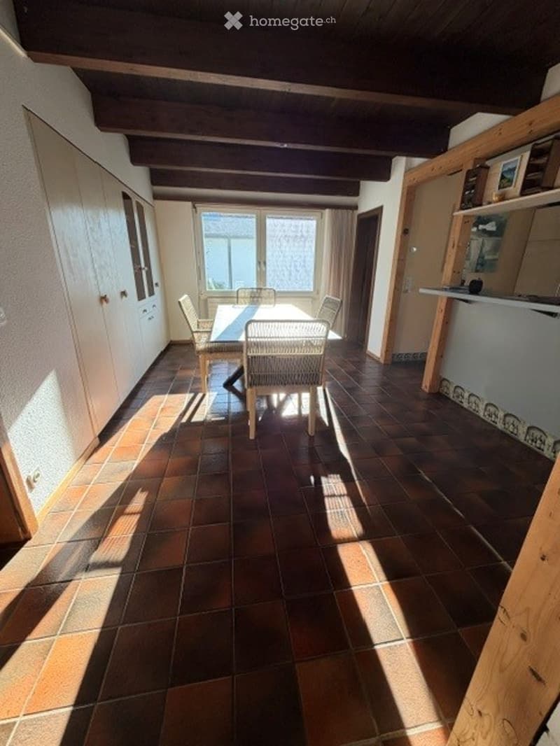 Dining area with a round wooden table and four chairs, large window with curtains, exposed wooden ceiling beams, terracotta tiled floor.