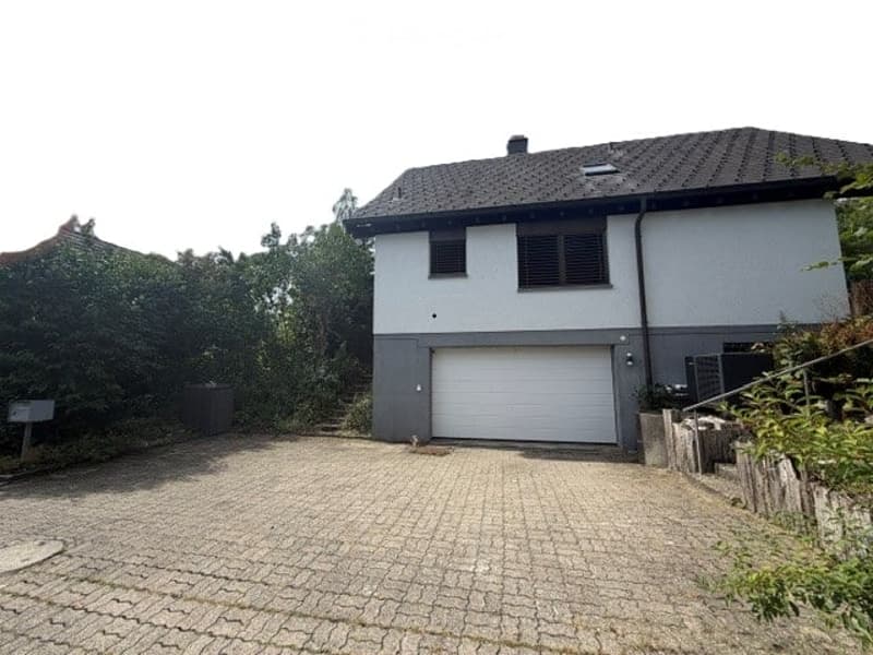 Two-story house with a gray garage door, surrounded by greenery and a paved driveway.