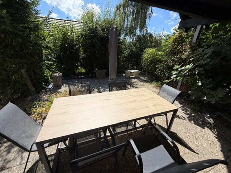 Outdoor patio area with a wooden table and four chairs, surrounded by greenery and trees.