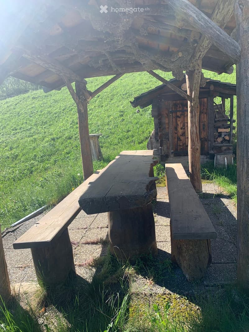 Wooden benches and table set under a wooden shelter with a rustic wooden cabin in the background, surrounded by green grass.