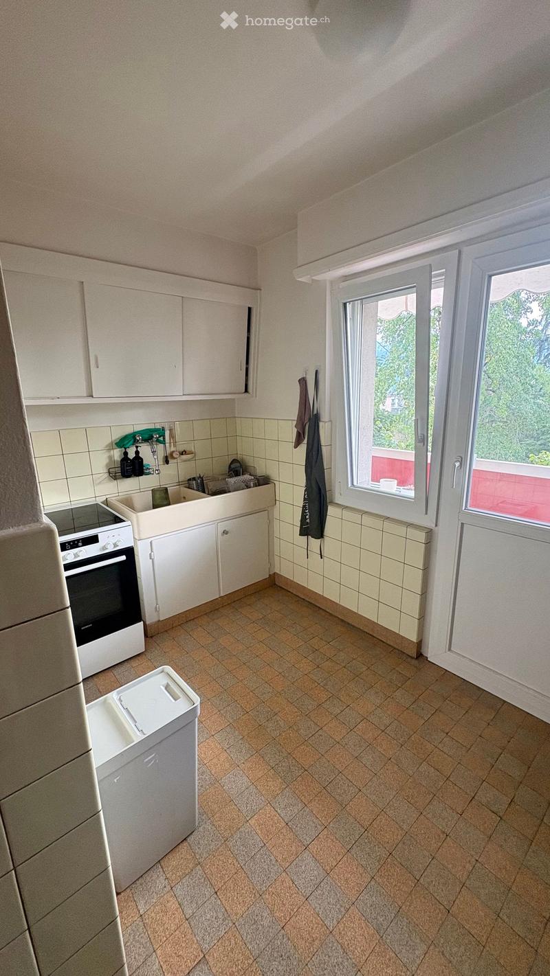 A small kitchen with white cabinets, a tiled backsplash, and a tiled floor. Features include a refrigerator, oven, sink, dishwasher, and a window with a view of greenery.