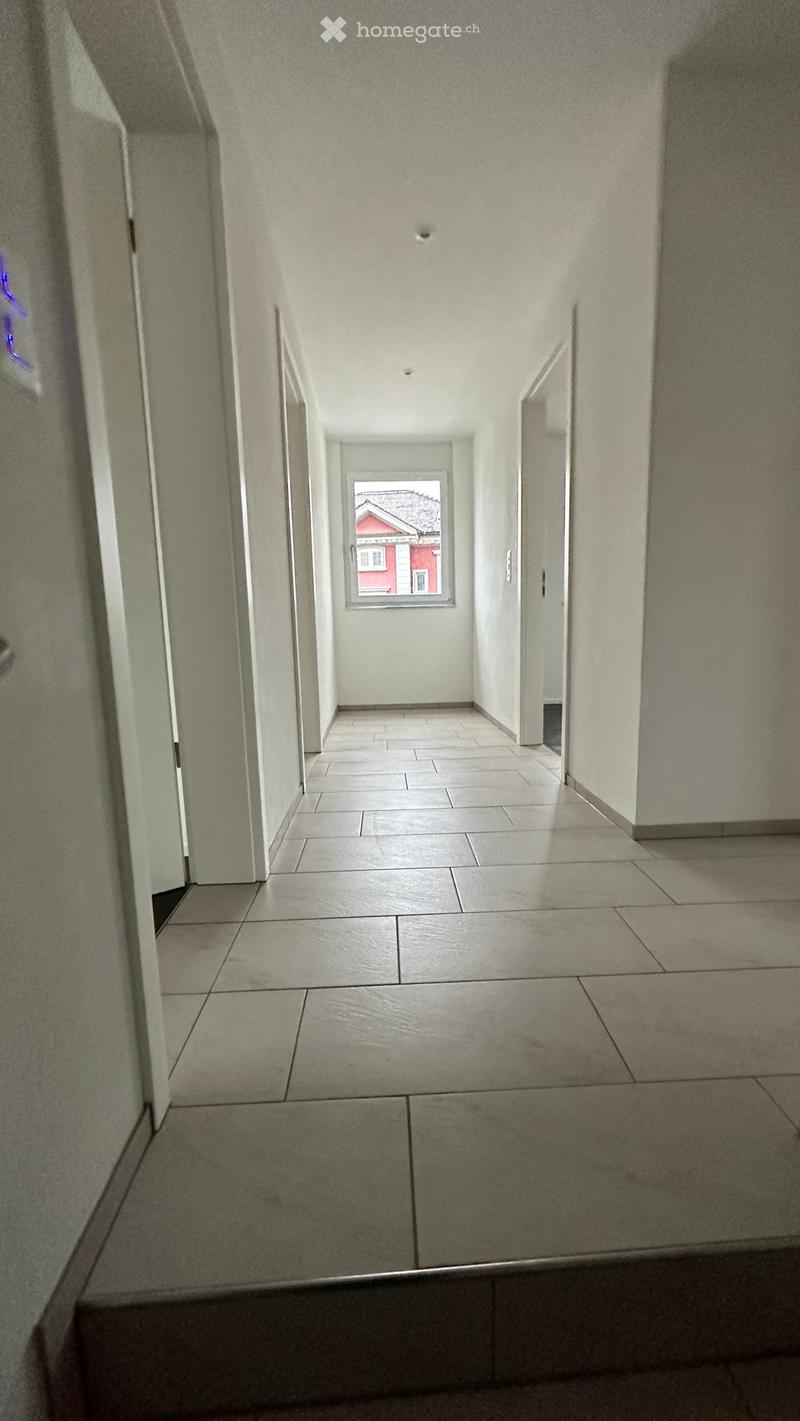 Empty hallway with tiled flooring, white walls, and a window at the end showing a red building outside.