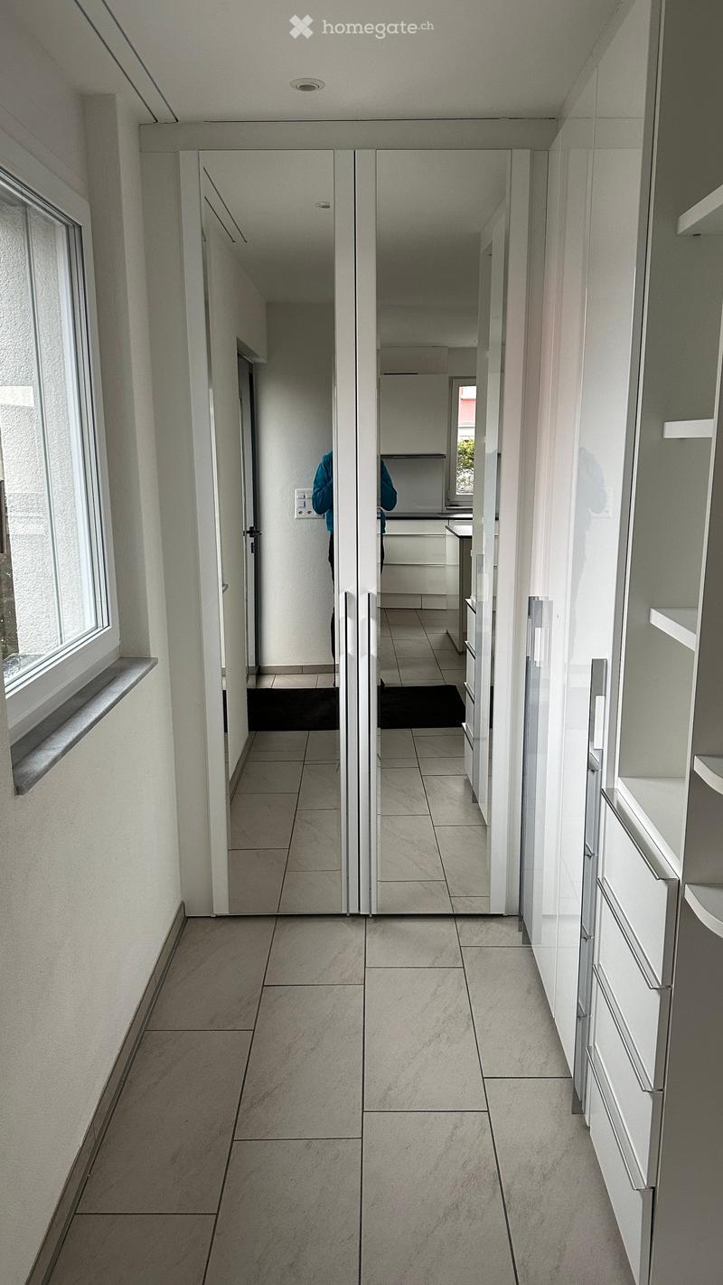 Modern hallway with tiled flooring, white walls, large windows, and mirrored doors leading to a kitchen area.