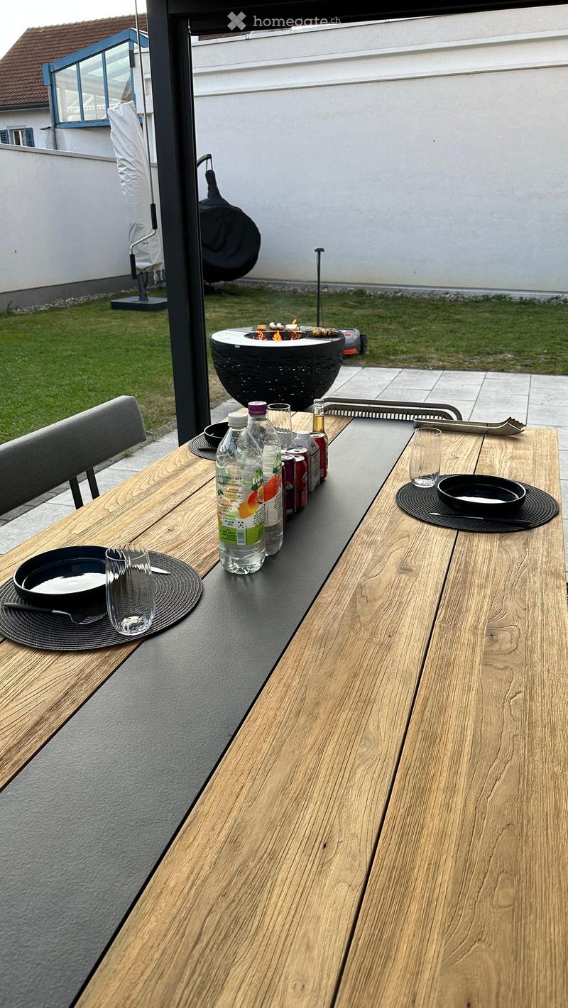A wooden outdoor table with a black surface, set with bowls, cups, and bottles, accompanied by a chair and a fire pit in the background.