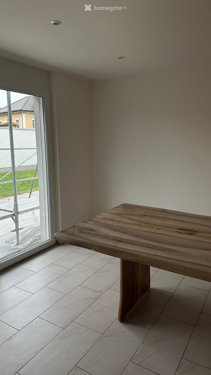 A modern, minimalist room with a large wooden table, light-colored tiled floor, and a large window with a view of the outside.
