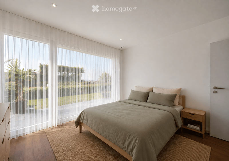 Modern bedroom with a large bed, nightstand, drawer, and a potted plant near the window. The room features wooden flooring, white walls, and sheer curtains allowing natural light to fill the space.