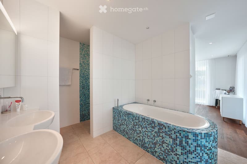 Modern bathroom with a large bathtub featuring blue mosaic tiles, two sinks, and a towel hanging on a rack. The space is bright with white tiles and recessed lighting.