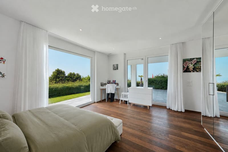 Bright bedroom with wooden flooring, large windows, and a view of greenery. Features a bed, nightstand, chair, desk, and mirror.