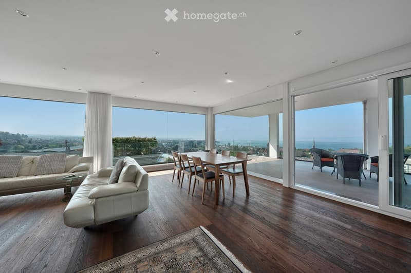Modern living and dining area with large windows offering a scenic view, featuring a white sectional sofa, a wooden dining table with chairs, and a patterned rug on a wooden floor.