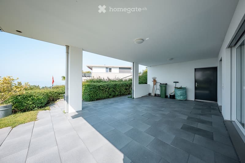 Spacious balcony with tiled flooring, white walls, and a covered roof. Features a door leading inside, a green trash bin, and some outdoor storage items.