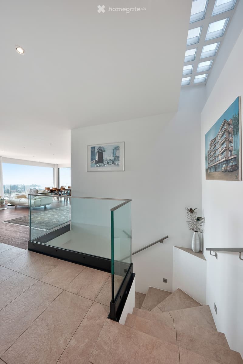 Modern hallway with a glass railing staircase, white walls, and a view of a living area with a sofa, dining table, and chairs.