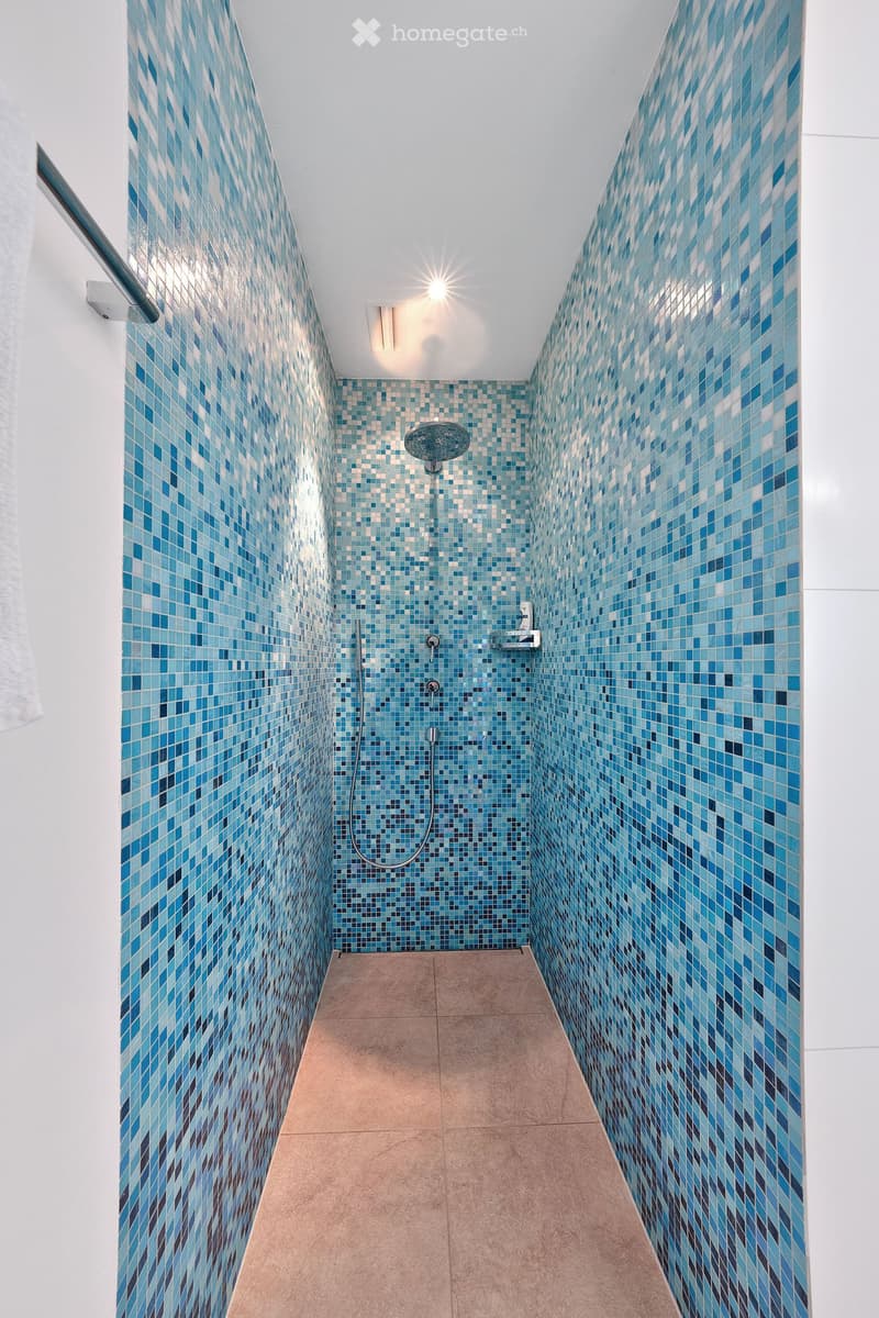 Modern shower area with blue mosaic tiles on walls, beige tiled floor, handheld showerhead, and built-in handrail.