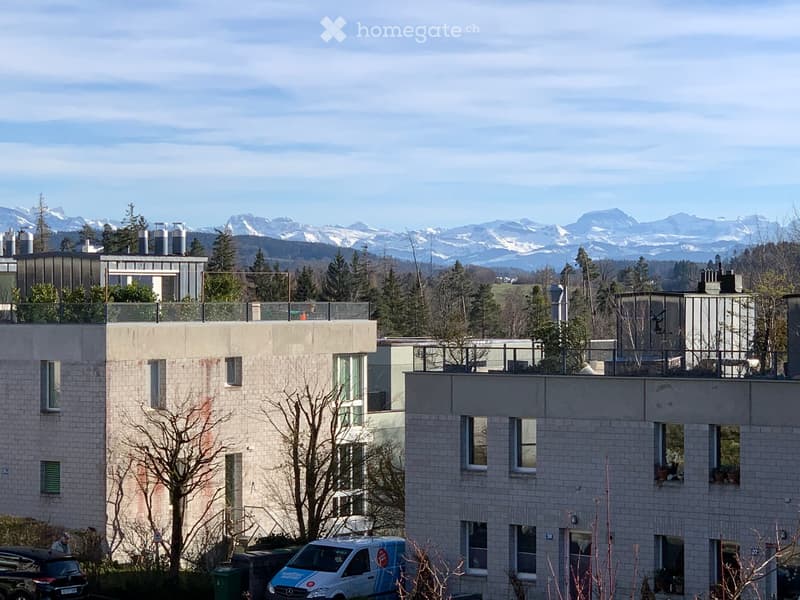 Modern multi-story residential building with balconies, surrounded by trees and mountains in the background.