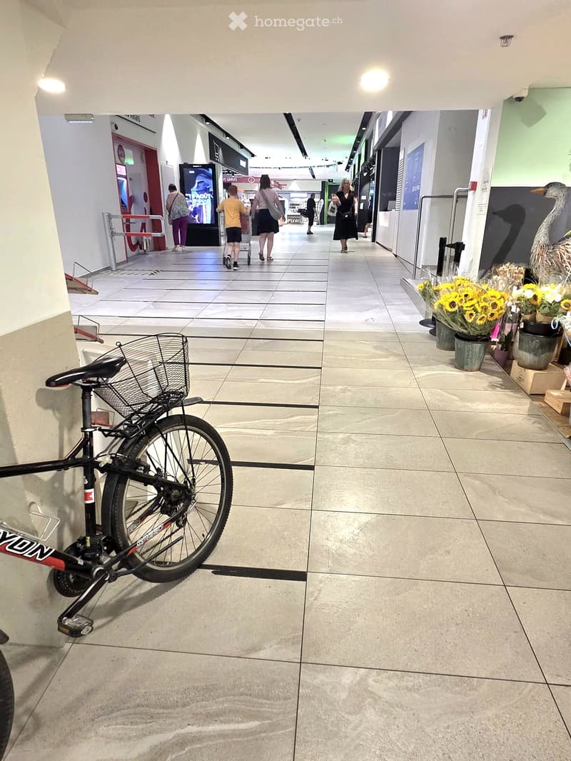 A spacious, well-lit hallway with tiled flooring, modern lighting, and a bicycle parked on the left side. The hallway features a large mirror reflecting the scene, with people walking in the distance and decorative flower arrangements on the right side.