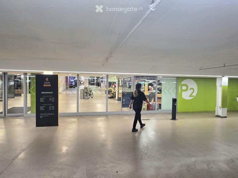 A spacious, modern hallway with polished concrete floors, large glass doors leading to a shopping area, and a sign welcoming visitors.