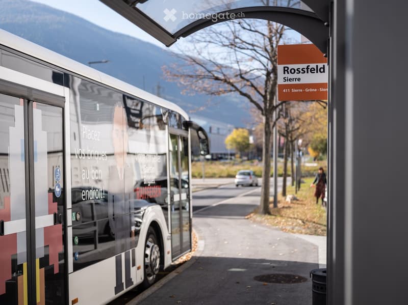A modern bus is parked at a bus stop with a sign reading "Rossfeld Sierre." The background shows a scenic mountain view and a few trees.