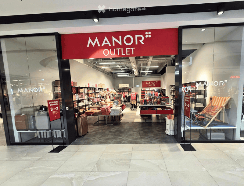 Storefront of Manor Outlet with glass windows displaying various items including luggage, shelves with folded towels, and a lounge chair.
