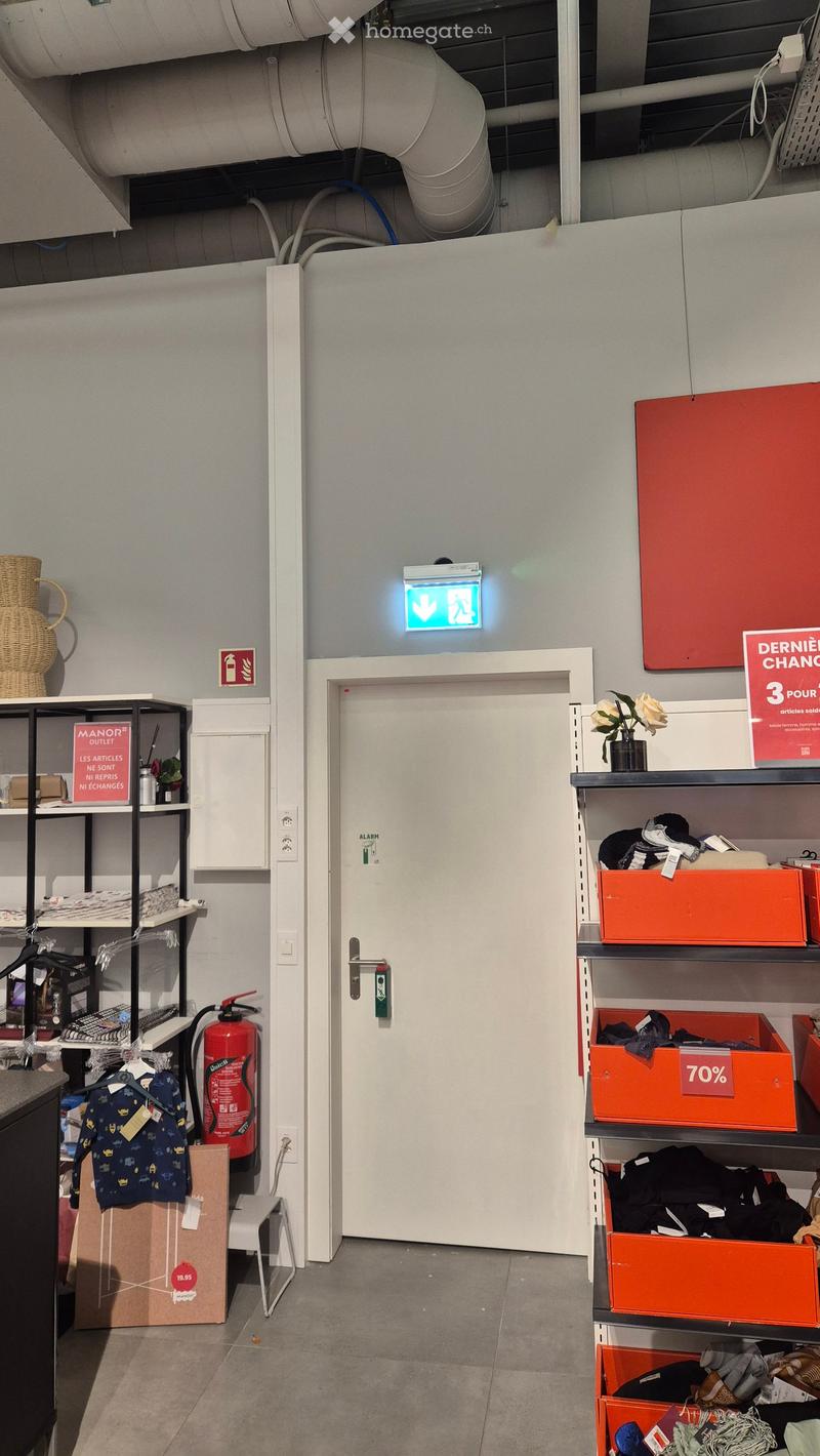 A well-lit hallway with a white door, shelves on both sides holding various items including clothing, boxes, and a fire extinguisher. A sign indicating an exit is visible above the door.