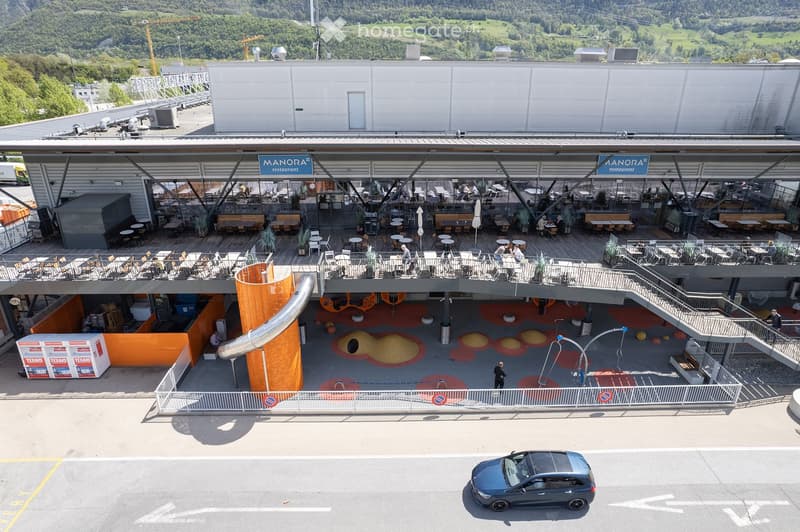 A modern building with a large outdoor seating area, featuring tables, chairs, and a restaurant sign. The area includes a colorful play area with a slide and other play structures.