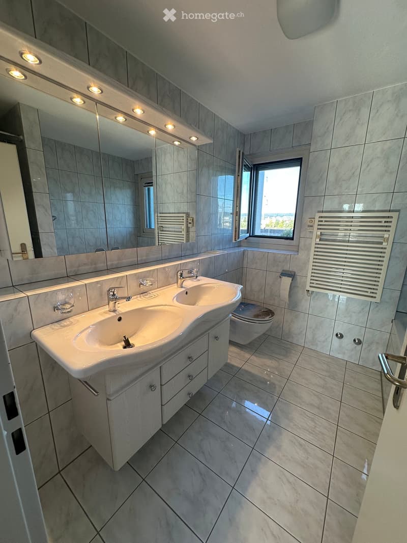 Modern bathroom with double sink, large mirror, toilet, radiator, and a window letting in natural light.