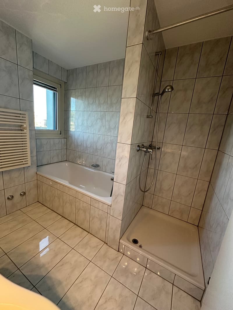 Modern bathroom with a bathtub, separate shower area, tiled walls and floor, and a window providing natural light.