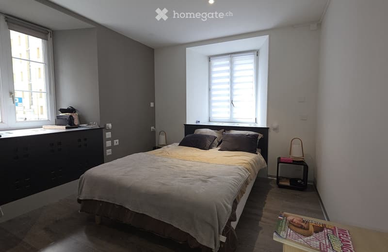 A modern bedroom with a large bed, nightstand, and a small table with a book on it. The room features wooden flooring, large windows with blinds, and a dark-colored wardrobe.