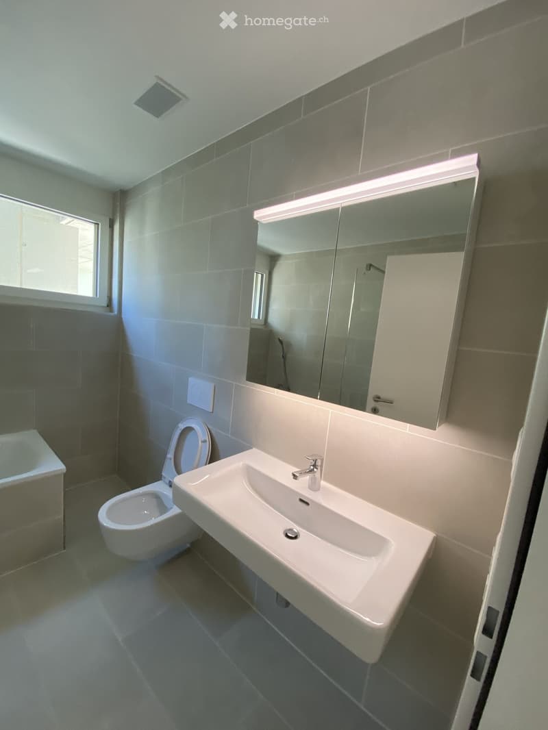 Modern bathroom with a white sink, chrome faucet, toilet, large mirror with built-in lighting, and a small window providing natural light.