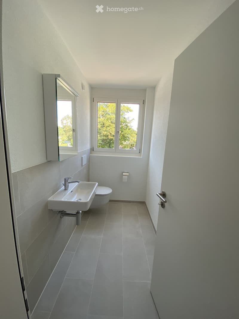Modern bathroom with white tiles, a sink, toilet, and a large window offering a view of greenery.