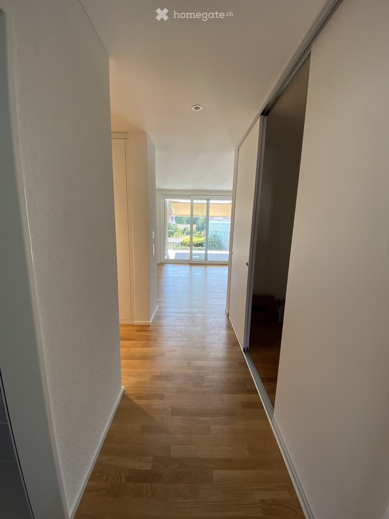 A modern hallway with wooden flooring, white walls, and recessed ceiling lighting. The hallway leads to a sliding glass door that opens to a balcony or outdoor area.
