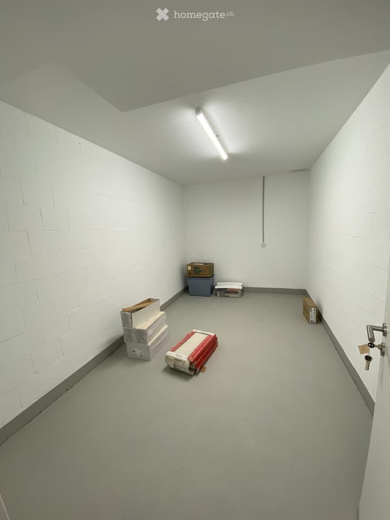 Empty room with white brick walls, grey floor, and fluorescent lighting. Several boxes and a small container are placed against the walls.