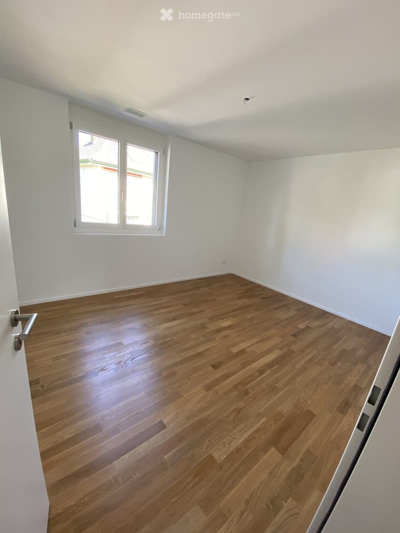 Empty room with light wooden flooring and white walls, featuring a large window with white frame and a door handle visible on the left side.