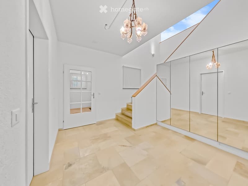 Bright, modern hallway with white walls, beige tiled floor, large mirror, staircase with wooden railing, and skylight ceiling.