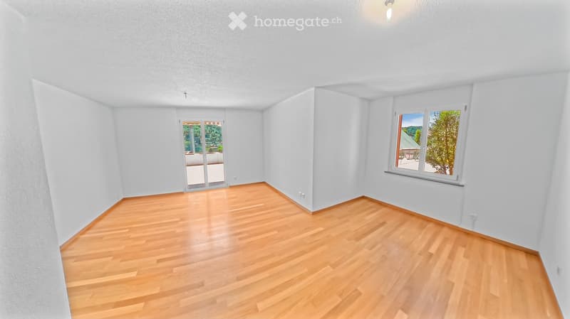 Empty room with light wooden flooring, white walls, and large windows offering a view of greenery outside. The room features a sliding glass door leading to a balcony or outdoor area.