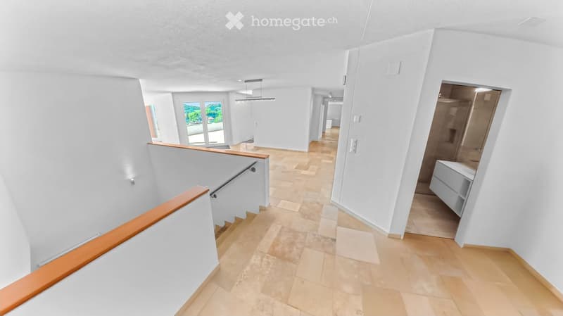 Bright, modern hallway with white walls, wooden flooring, and a staircase leading upwards. Features include recessed lighting, a wooden railing, and a view of a room with large windows and a balcony.