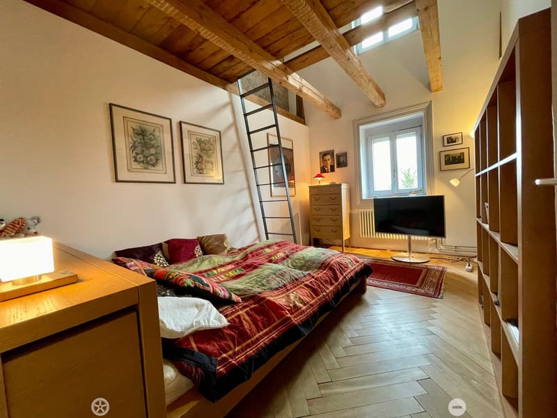 Cozy bedroom with wooden floors, slanted wooden ceiling beams, a large window, a bed with colorful quilt, nightstand with lamp, TV, dresser, and picture frames on the walls.