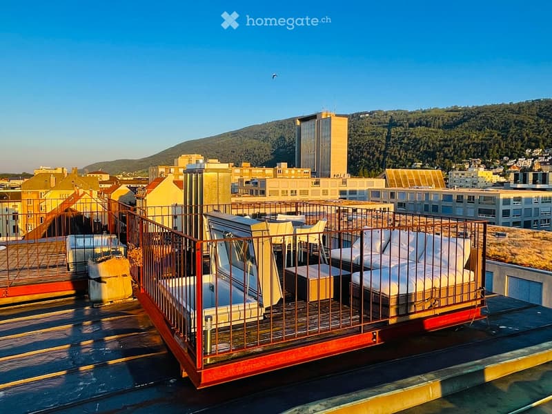 Rooftop terrace with a sofa, chairs, and a table, overlooking a cityscape with buildings and hills.