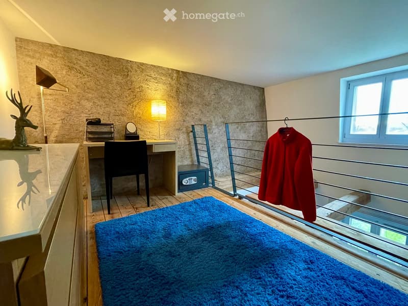 A furnished room with a desk, chair, lamp, mirror, security box, a red coat hanging on a rack, and a decorative deer statue on a shelf.