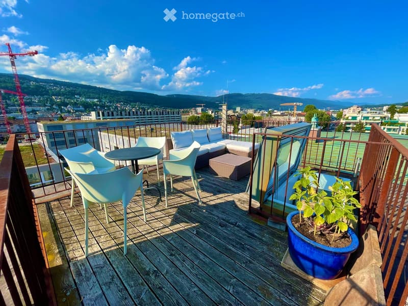 A wooden balcony with white furniture including a sofa, chairs, a small table, and a potted plant, overlooking a cityscape with mountains in the background.