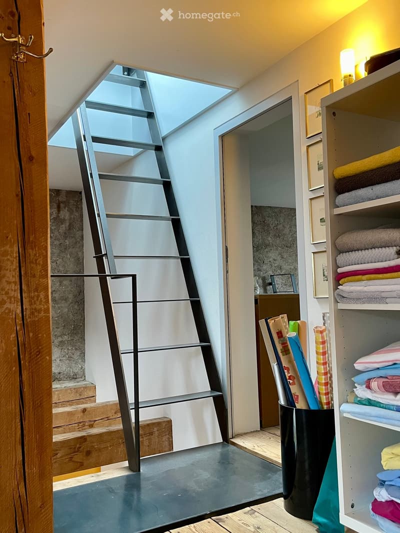 Modern, minimalist room with a sleek staircase leading upwards, a shelf with neatly folded towels, framed pictures, and a storage bin holding rolled materials.