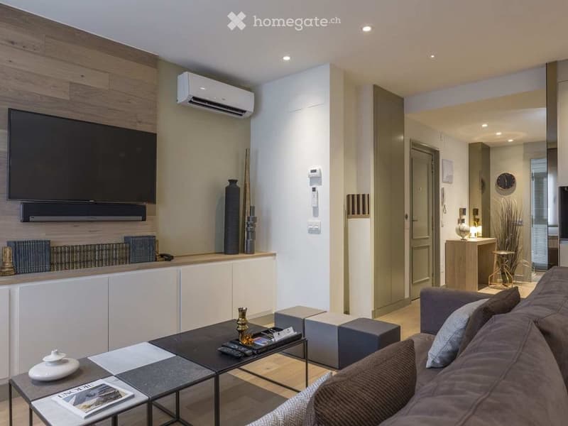 Modern living room with a grey sofa, coffee table, decorative vases, and a television mounted on a wooden wall. The space features built-in cabinets, a radiator, and a sleek design with warm lighting.