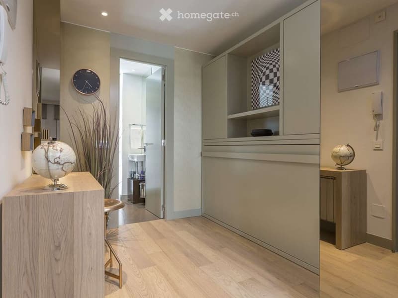 Modern hallway with light wooden flooring, a small wooden stool, a globe on a table, a wall-mounted clock, and an open doorway leading to a bathroom with a sink and mirror.